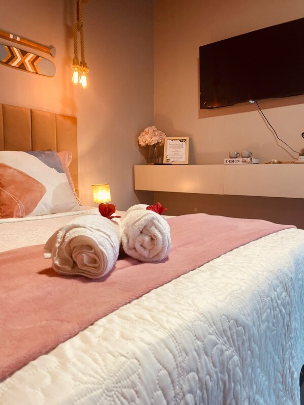 Un lit bien fait, avec du linge de lit blanc et rose, un luminaire décoratif fixé au mur, un téléviseur à écran plat et une petite étagère avec une carte encadrée et deux tasses.