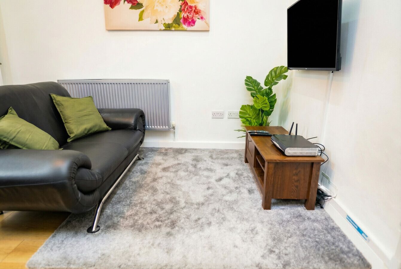 A living room with a black leather sofa, a grey carpet, a wooden side table with a laptop, and a large plant.