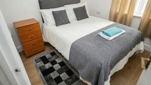 A bedroom with a bed, a wooden nightstand, a checkered rug, and a window with curtains.