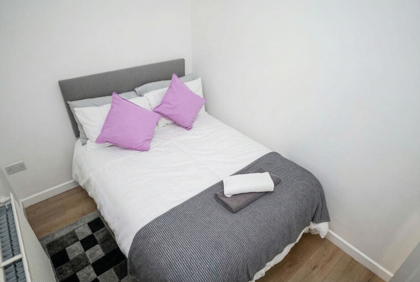 Uma cama bem arrumada com cabeceira cinzenta, roupa de cama branca e dois travesseiros roxos.