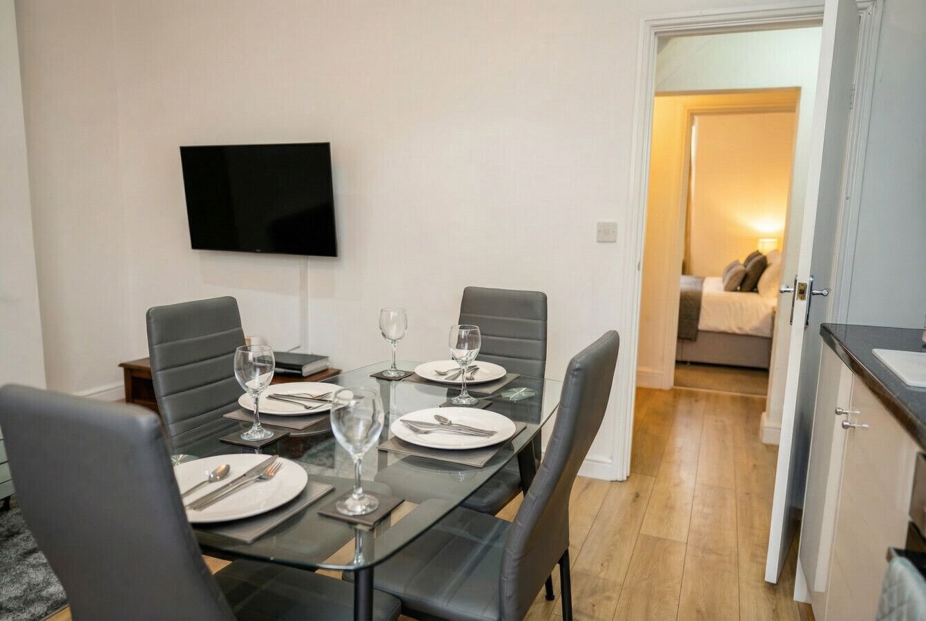Un comedor con una mesa dispuesta para cuatro, un televisor de pantalla plana montado en la pared y un acceso a un dormitorio.