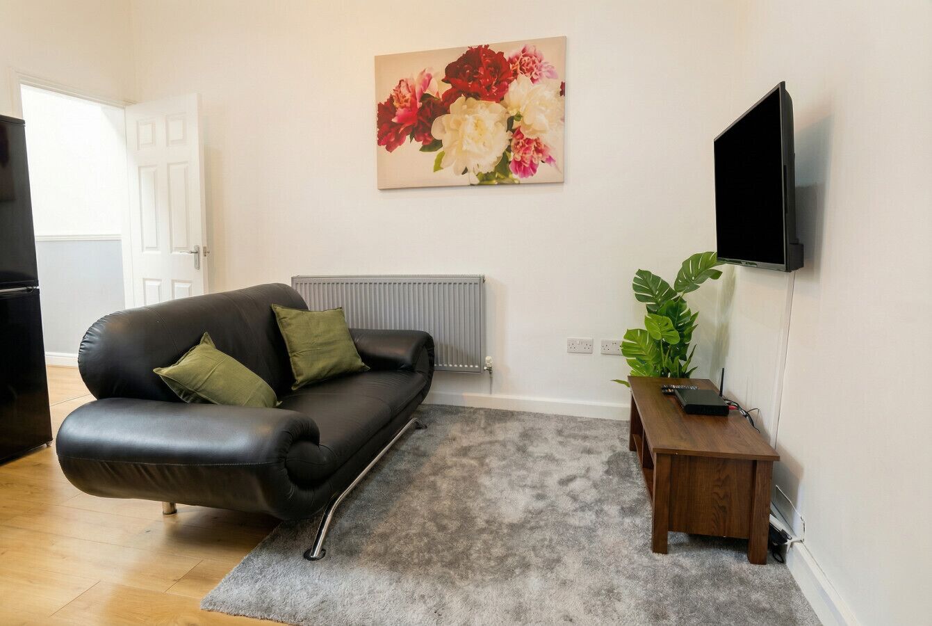 Una sala de estar con un sofá de cuero negro, una alfombra gris, una mesita de madera, un televisor montado y un cuadro floral en la pared.