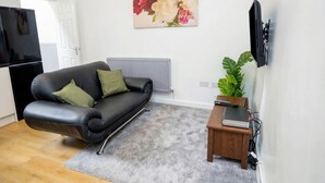 A living room with a black leather sofa, a grey carpet, a wooden side table with a laptop, a wall-mounted TV, and a floral painting.