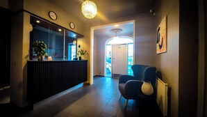 A reception area with a dark wooden counter, a potted plant, and a wall clock. There is a chair with a textured cushion and a framed picture on the wall. - Nupan Deluxe  (Keflavík)