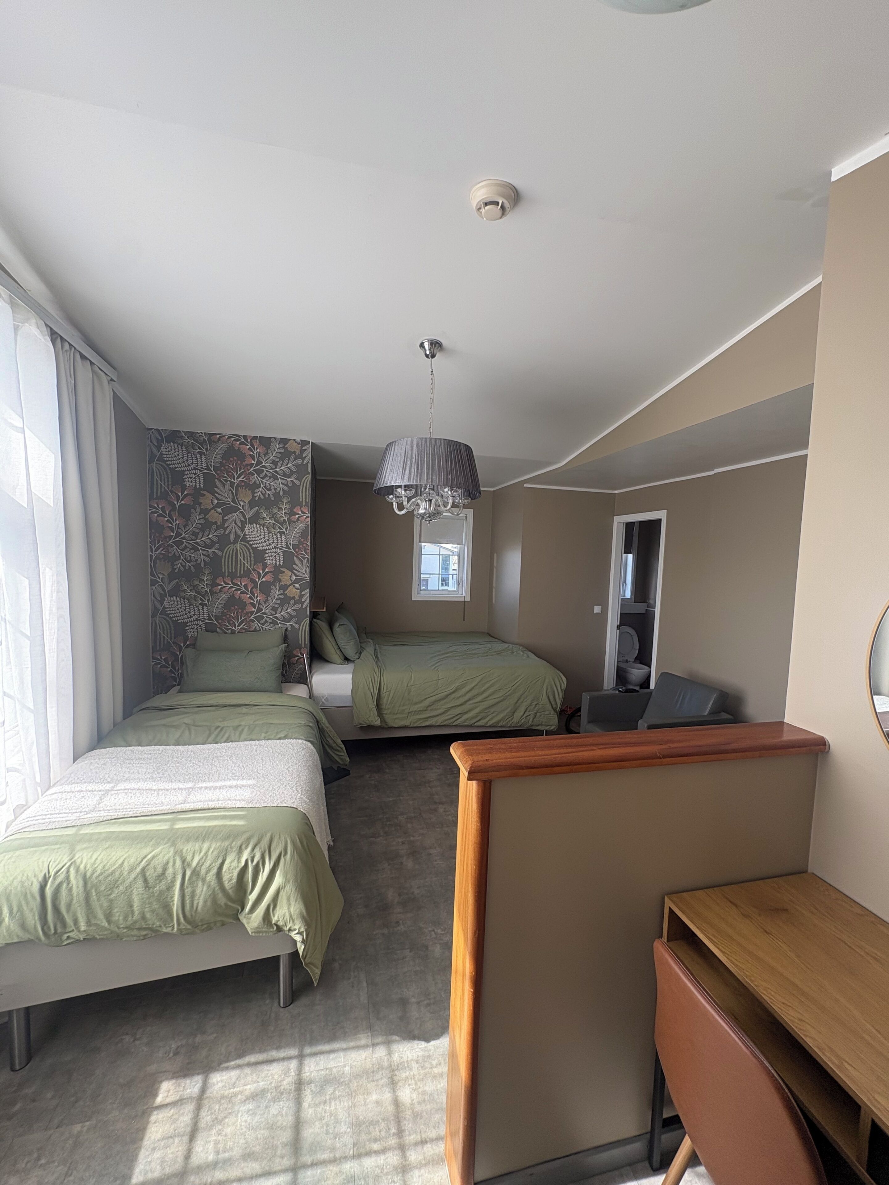 A hotel room with two beds, a desk, a chair, a hanging lamp, and a window with curtains.
