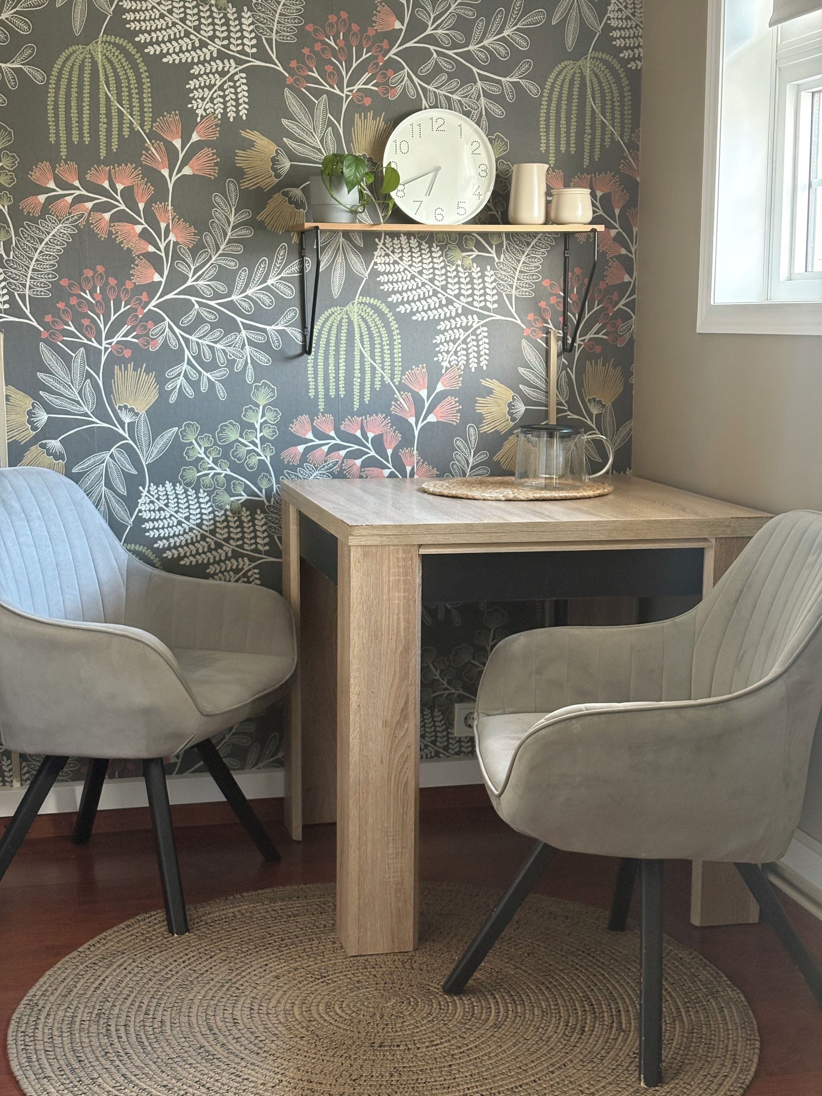 Una habitación con una mesa de madera, dos sillas, un reloj, una estantería con objetos decorativos y un papel tapiz estampado.