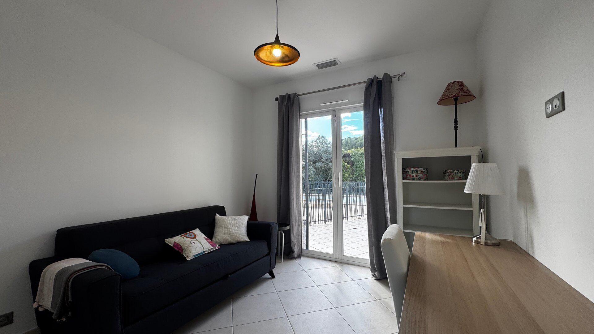 Un salón moderno con un sofá negro, una mesa de centro de madera, una lámpara y vistas a través de un ventanal corredero.