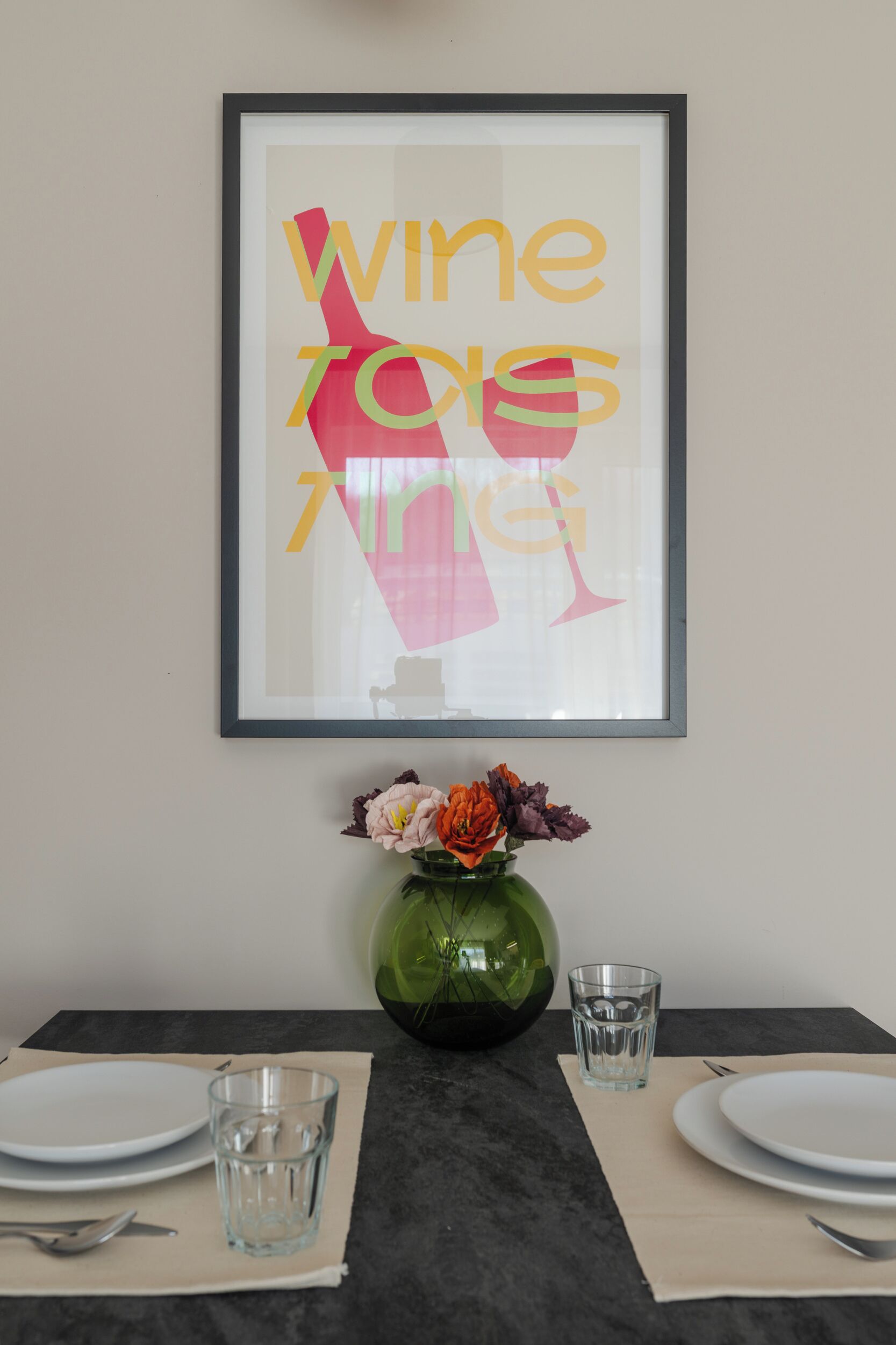 A framed artwork with the text "wine to taste" on a wall, a vase with flowers, and a table set with plates, cutlery, and glasses.