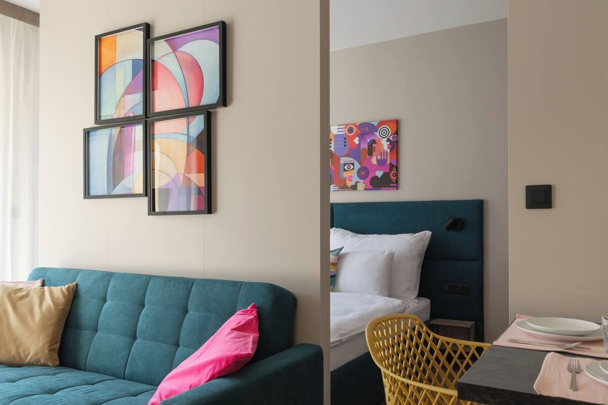 Una habitación de hotel moderna con un sofá azul verdoso, un sillón amarillo, una cama con cabecera azul y arte abstracto en las paredes.