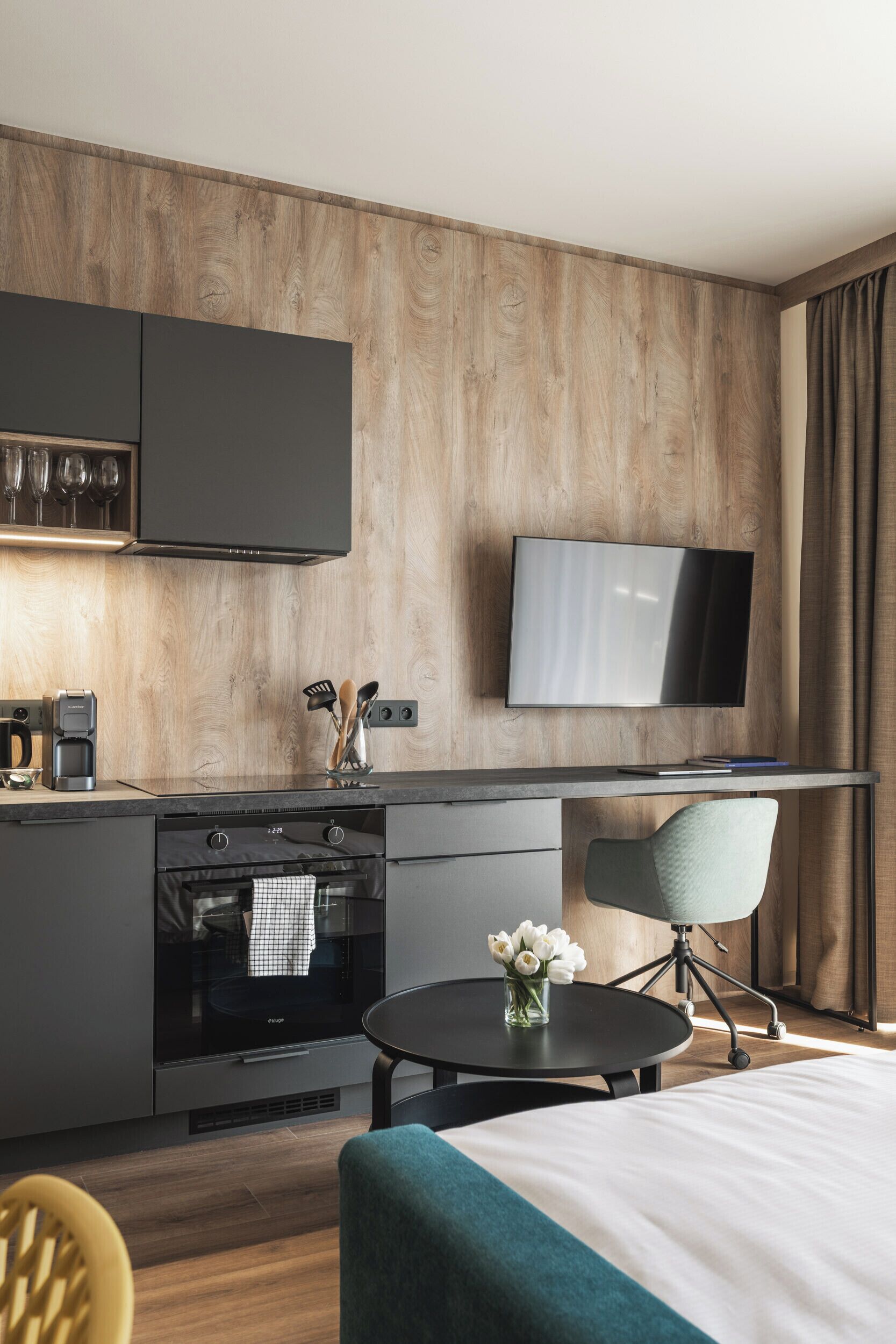 A modern hotel room with a wooden accent wall, a flat-screen TV, a kitchenette, a dining area, and a bed.