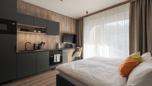 A modern hotel room with a kitchen area, a bed with pillows, and a large window with curtains.