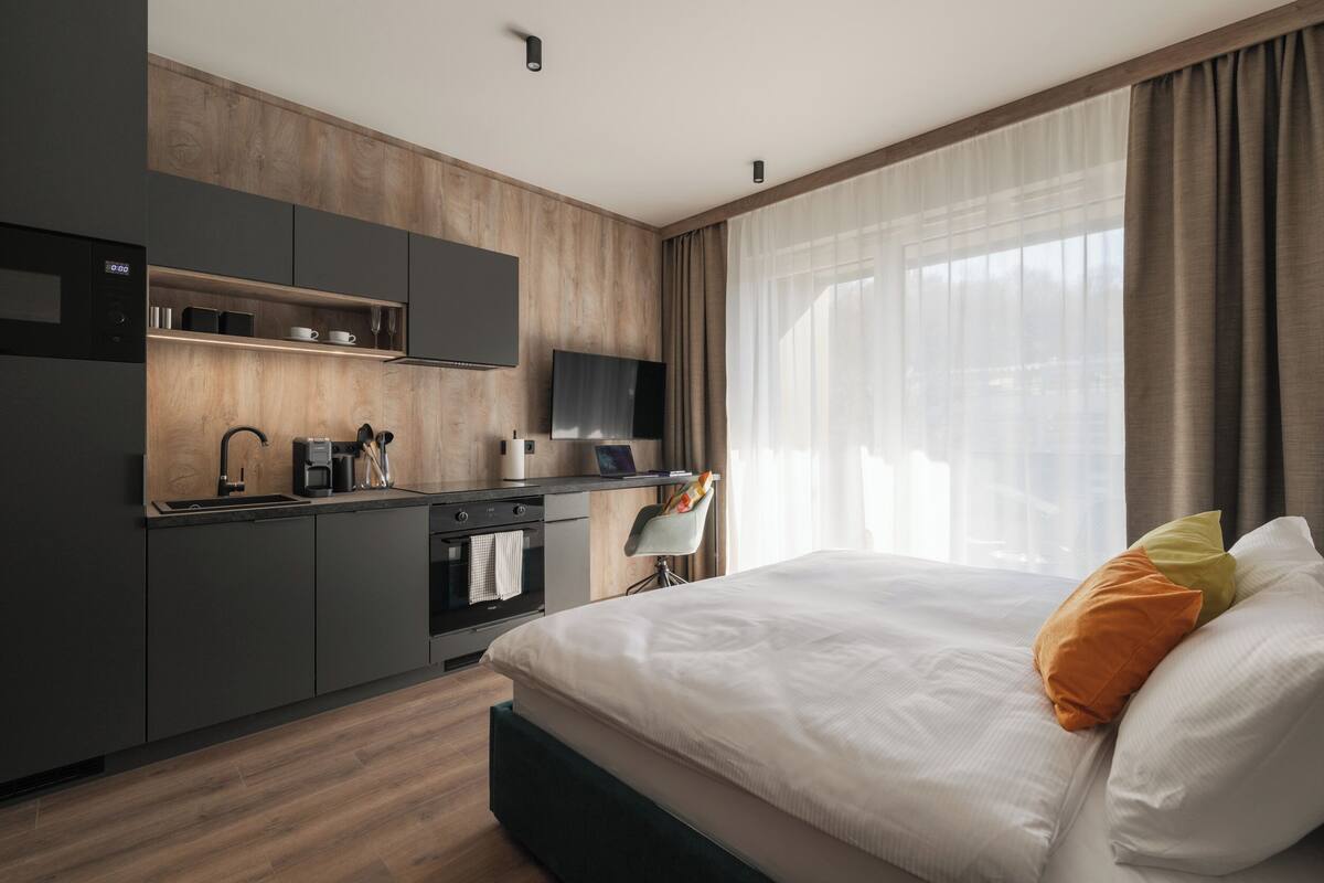 Una habitación de hotel moderna con cocina, cama con almohadas y un amplio ventanal con cortinas.