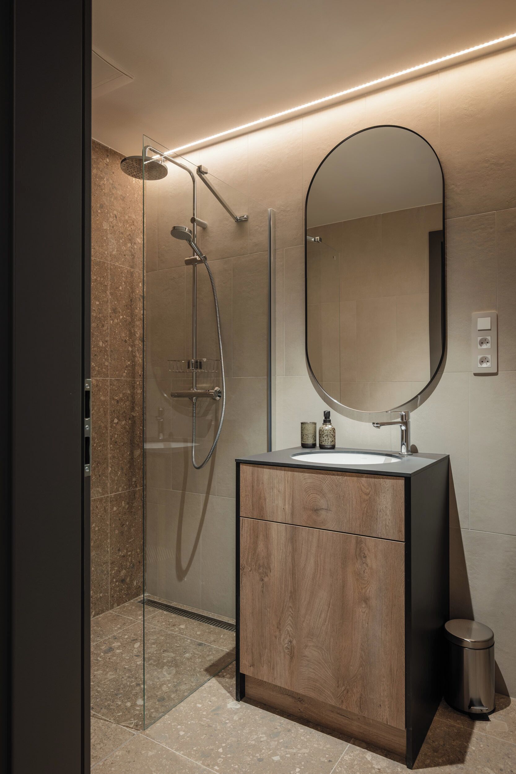 Un baño moderno con ducha envuelta en vidrio, tocador de madera, un espejo grande y un pequeño cubo de basura.