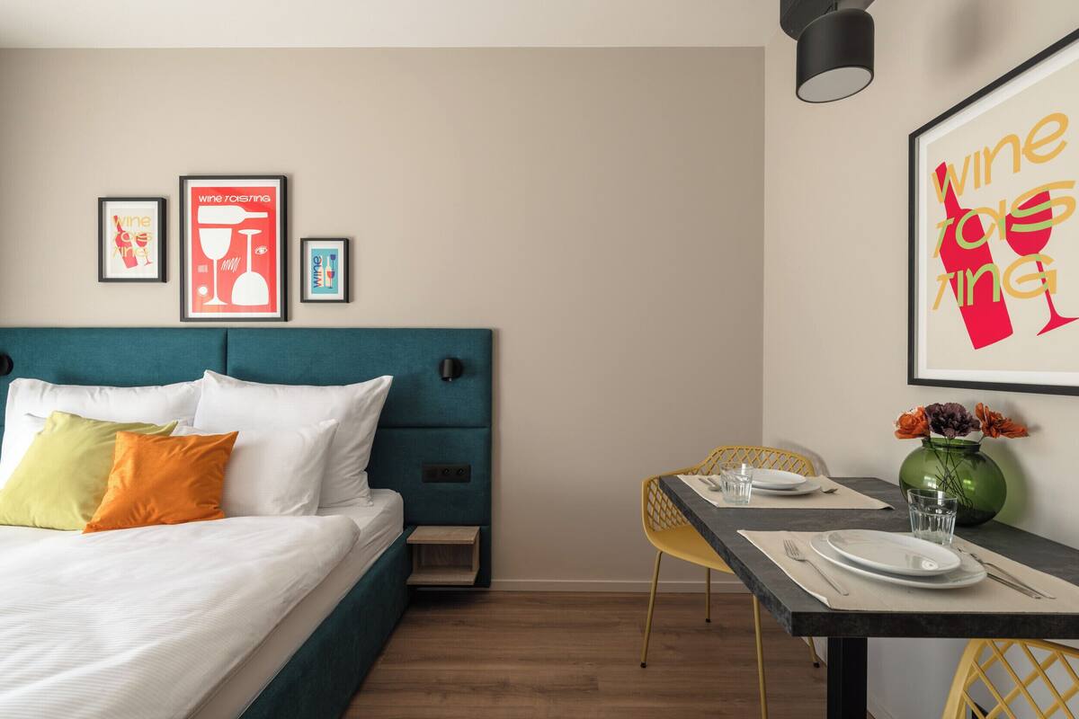 Una habitación de hotel moderna con una cama de cabecera color turquesa, ropa de cama blanca y almohadas coloridas. Hay una zona de comedor con una silla amarilla, un jarrón verde con flores y un cartel enmarcados sobre el vino en la pared.