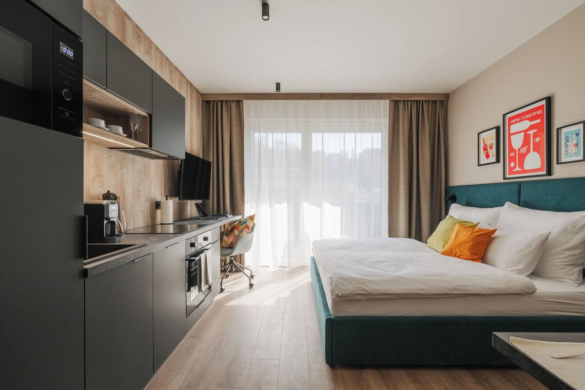 Una habitación de hotel moderna con kitchenette, una cama con almohadas y un gran ventanal con cortinas.