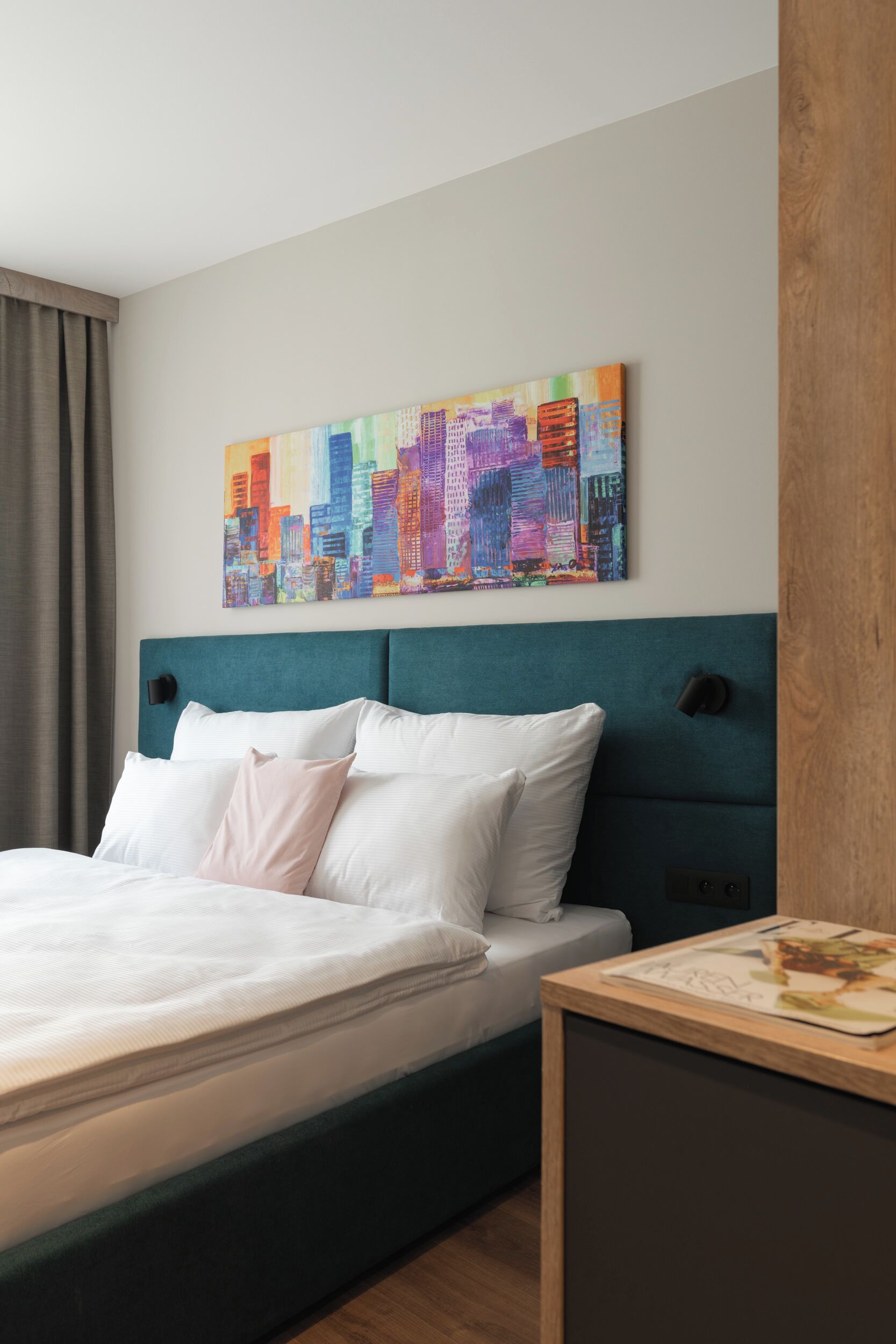 A neatly made bed with white linens and a teal headboard, a colorful cityscape painting on the wall, and a wooden nightstand with a magazine.