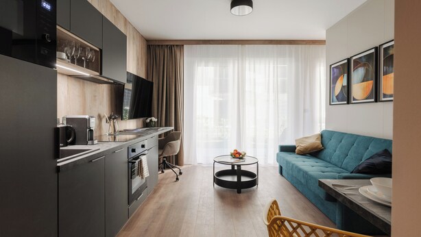 A modern kitchen with dark cabinetry, a built-in oven, and a coffee maker. There is a round dining table with a bowl of fruit and a chair. A teal sofa and a small table with a bowl are in the living area.