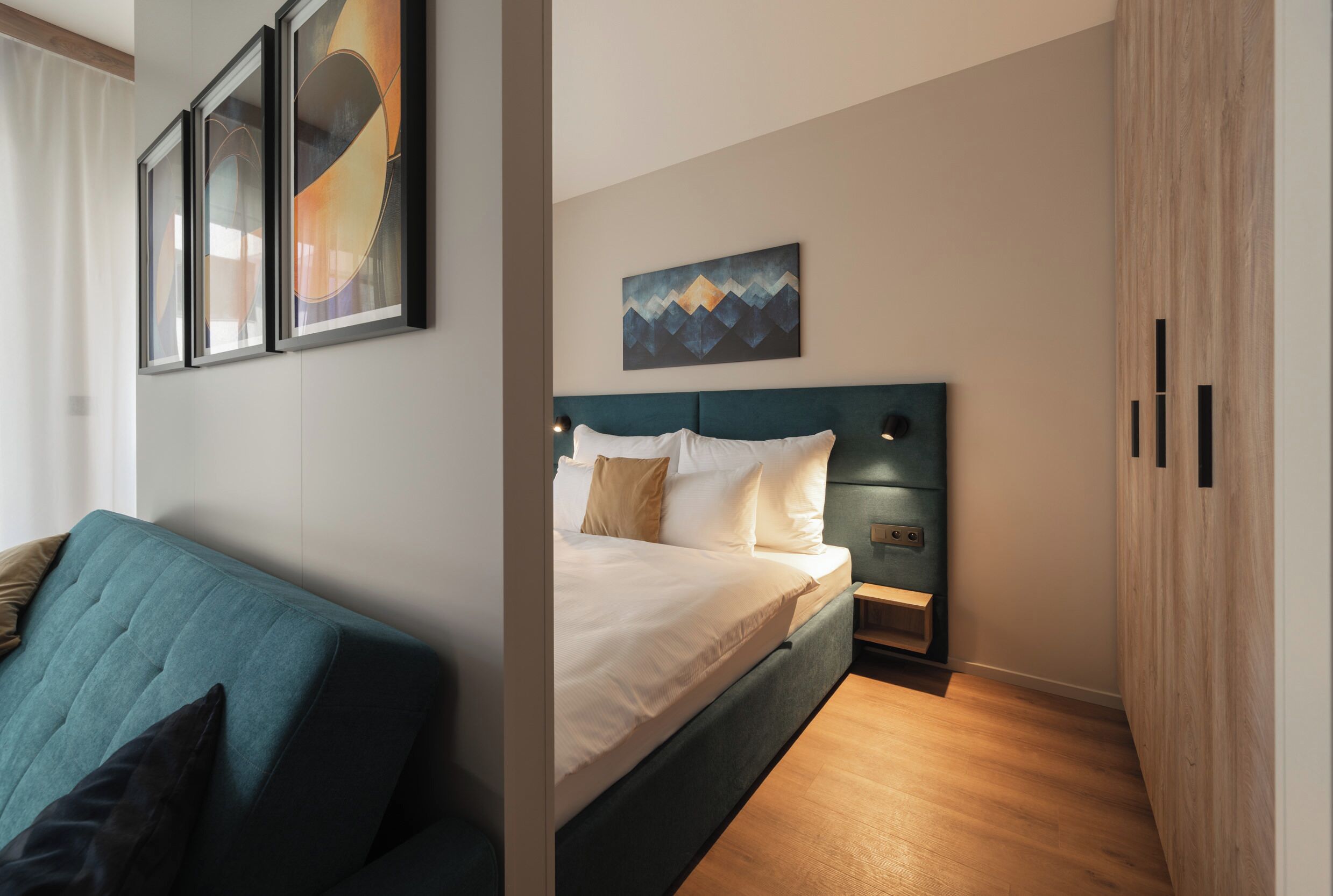 Una camera d'albergo moderna con un divano color petrolio, un letto rifatto con lenzuola bianche, un'opera d'arte appesa al muro e un'armadiatura in legno.