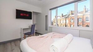 Ein modernes Hotelzimmer mit einem Bett, einem Schreibtisch mit Stuhl, einem an der Wand befestigten Fernseher und einem großen Fenster mit Blick auf Gebäude.