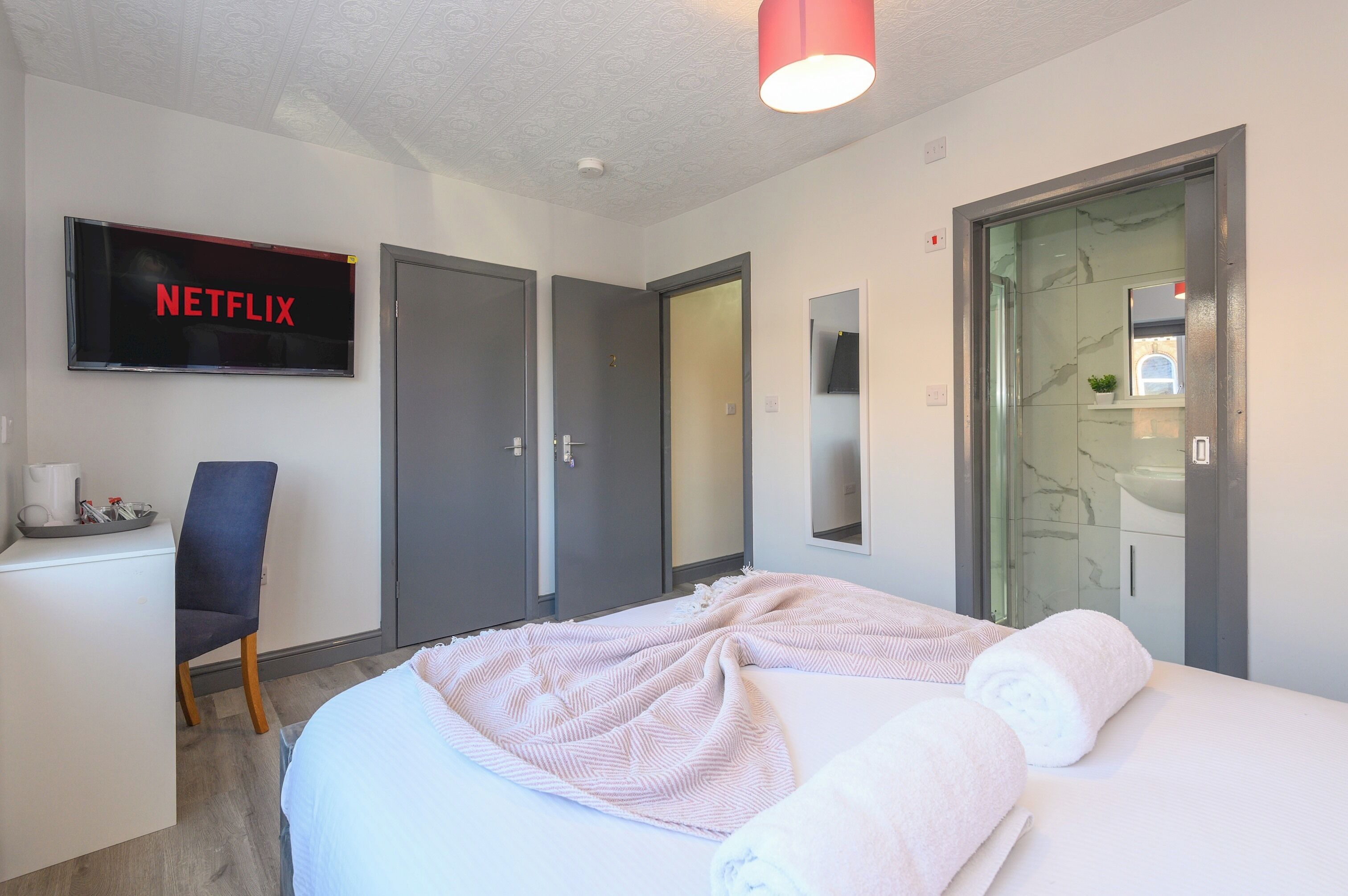 A hotel room with a bed, a desk, a chair, a TV displaying 'NETFLIX', and a bathroom with a glass shower.