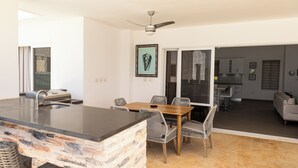Cocina moderna con una isla de piedra, comedor con sillas de mimbre y vista al salón.