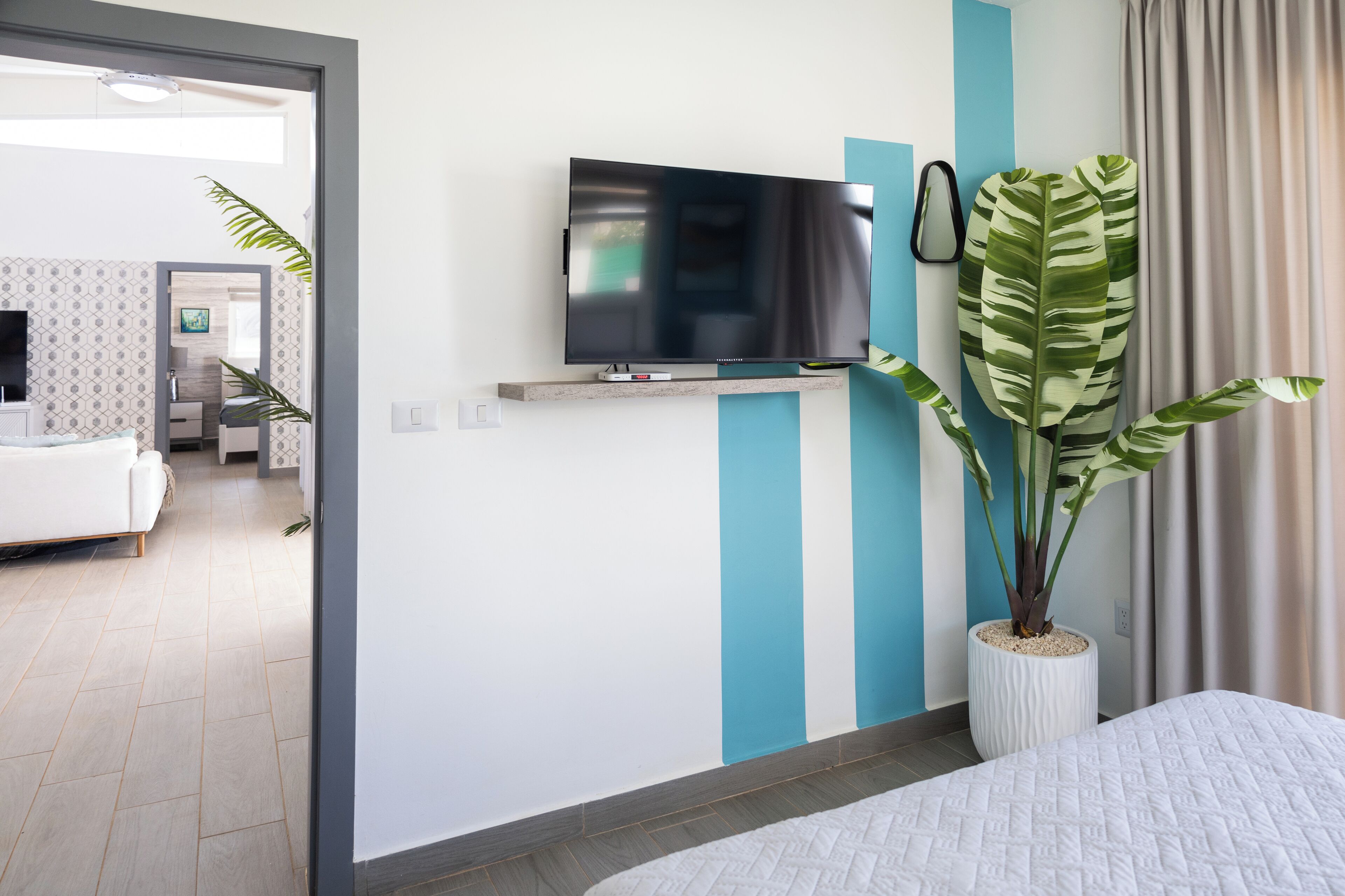Une chambre d’hôtel moderne dotée d’un téléviseur à écran plat fixé au mur, d’une grande plante en pot et d’un lit recouvert d’une courtepointe blanche.