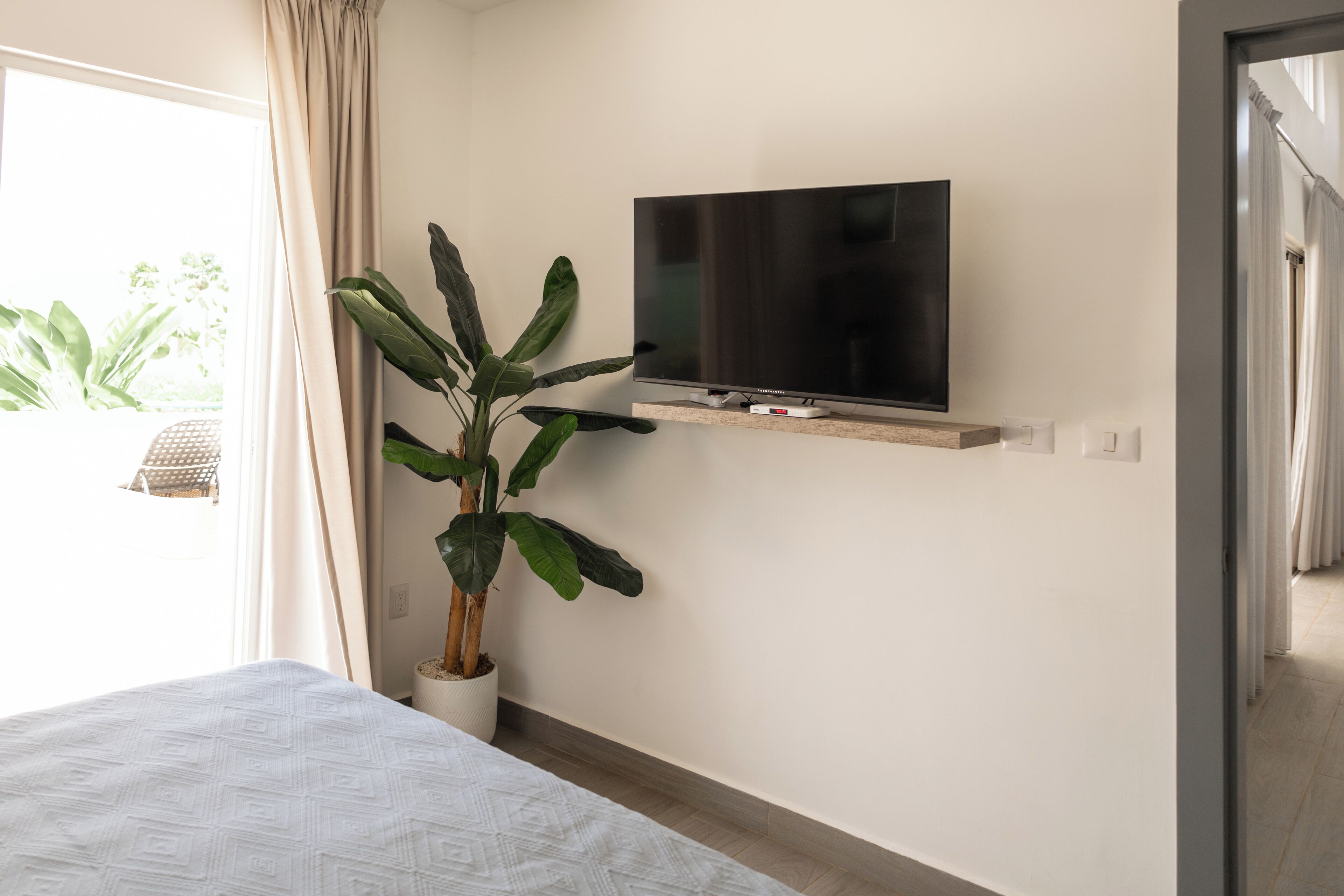 A bedroom with a bed, a potted plant, a TV on a shelf, and a door leading to a balcony.