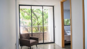 Une chambre moderne dotée d’une porte-fenêtre coulissante donnant sur un balcon, d’un fauteuil marron et d’un lit avec une tête de lit blanche.