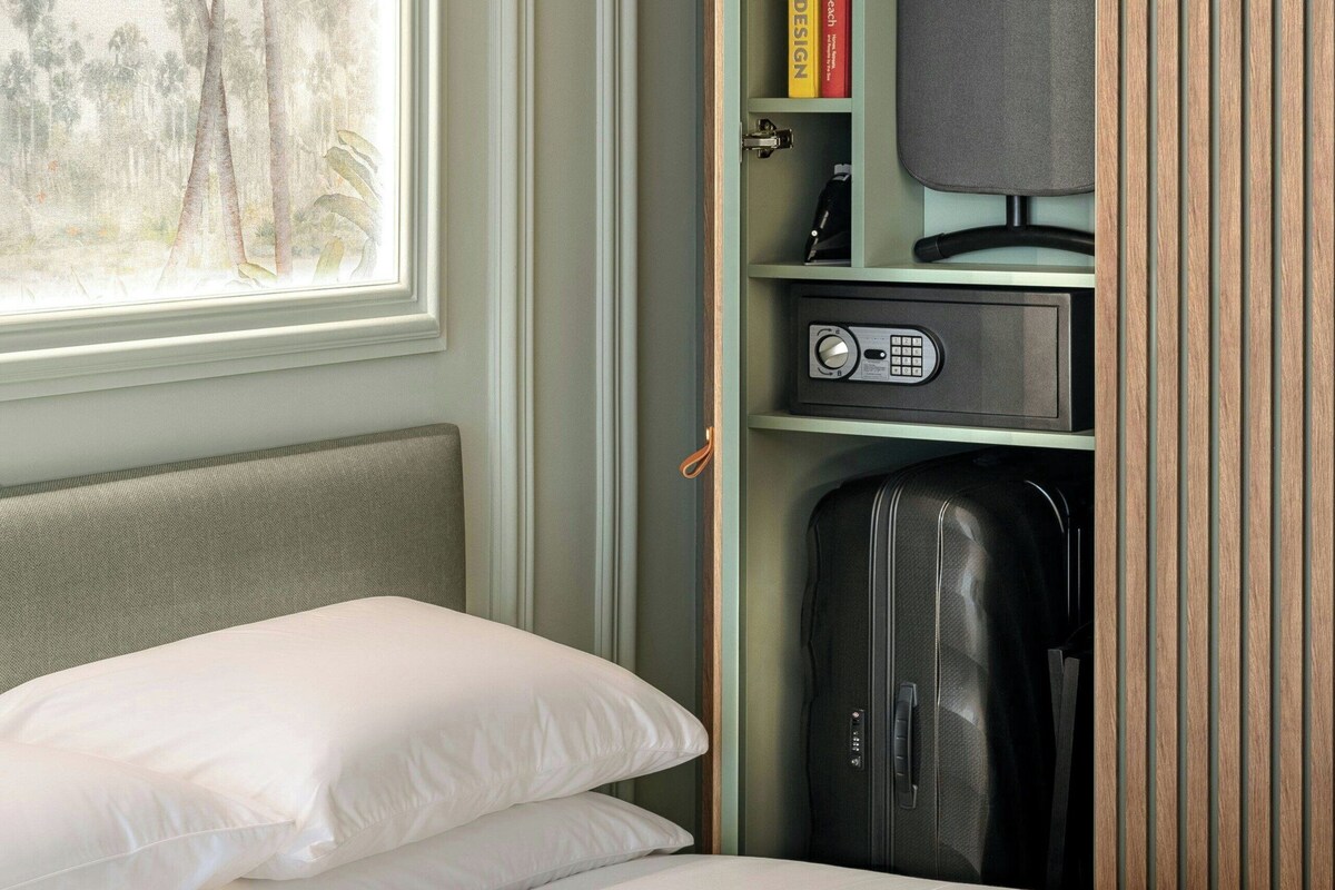 Ein Hotelzimmer mit Bett, Fenster mit Blick auf Palmen, Kleiderschrank mit Hängestuhl und Safe.