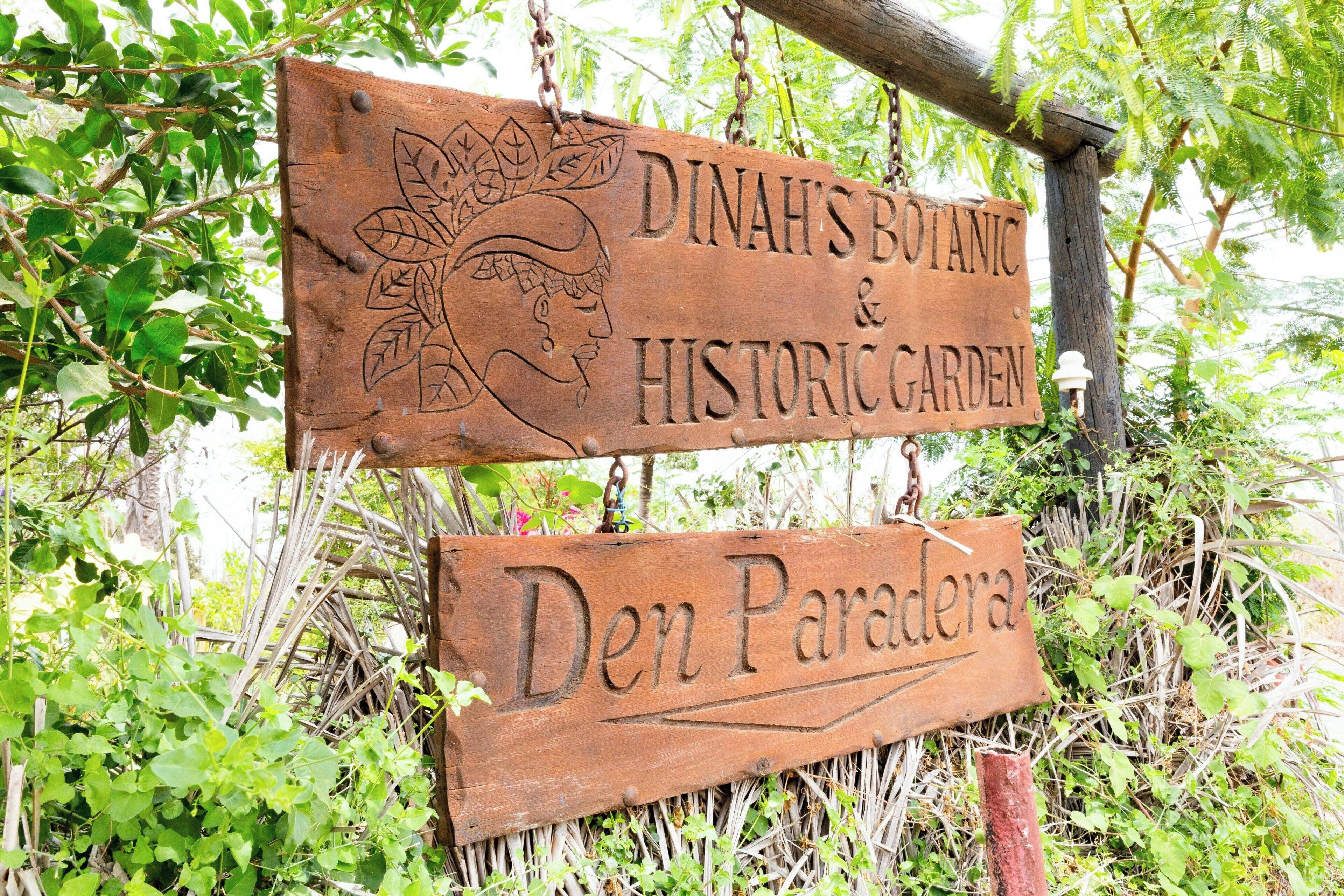 A wooden sign with carvings and text indicating 'Dina's Botanical & Historic Garden' and 'Den Paradera'.
