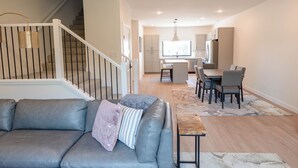 A modern living room with a gray sofa, a coffee table, and a dining area with chairs and a table. A staircase with a railing is visible in the background. - NashLo Boji Retreat (Arnolds Park)