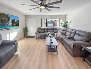 A spacious living room with a ceiling fan, a large flat-screen TV, and a view of a house through the open door. - Gather Together Garage Pets (Montrose)