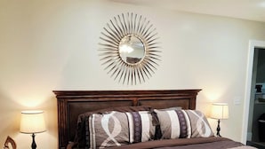 Una cama bien hecha con colcha estampada, dos lámparas de noche, un espejo en la pared y una pequeña mesita de madera con una lámpara.