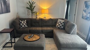 Una sala de estar con un sofá seccional, dos otomanas, una mesa de centro, una lámpara y un cuadro en la pared.