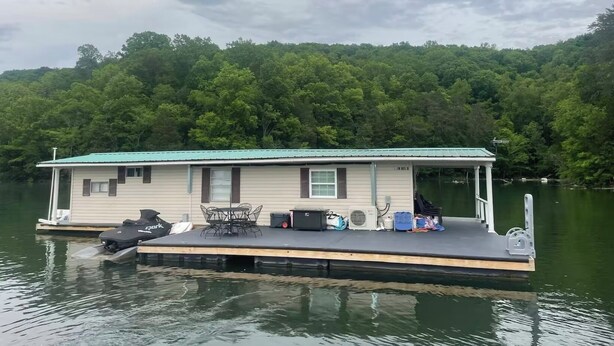 Una casa flotante con cubierta, una mesa, sillas y un motorboat amarrado al frente.