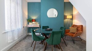Een eetruimte met een ronde tafel, groene stoelen, een klein bijzettafeltje met een lamp en een spiegel aan de muur.
