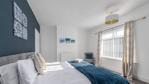 Een slaapkamer met een bed, nachtkastje, radiator, raam met gordijnen en wanddecoratie.