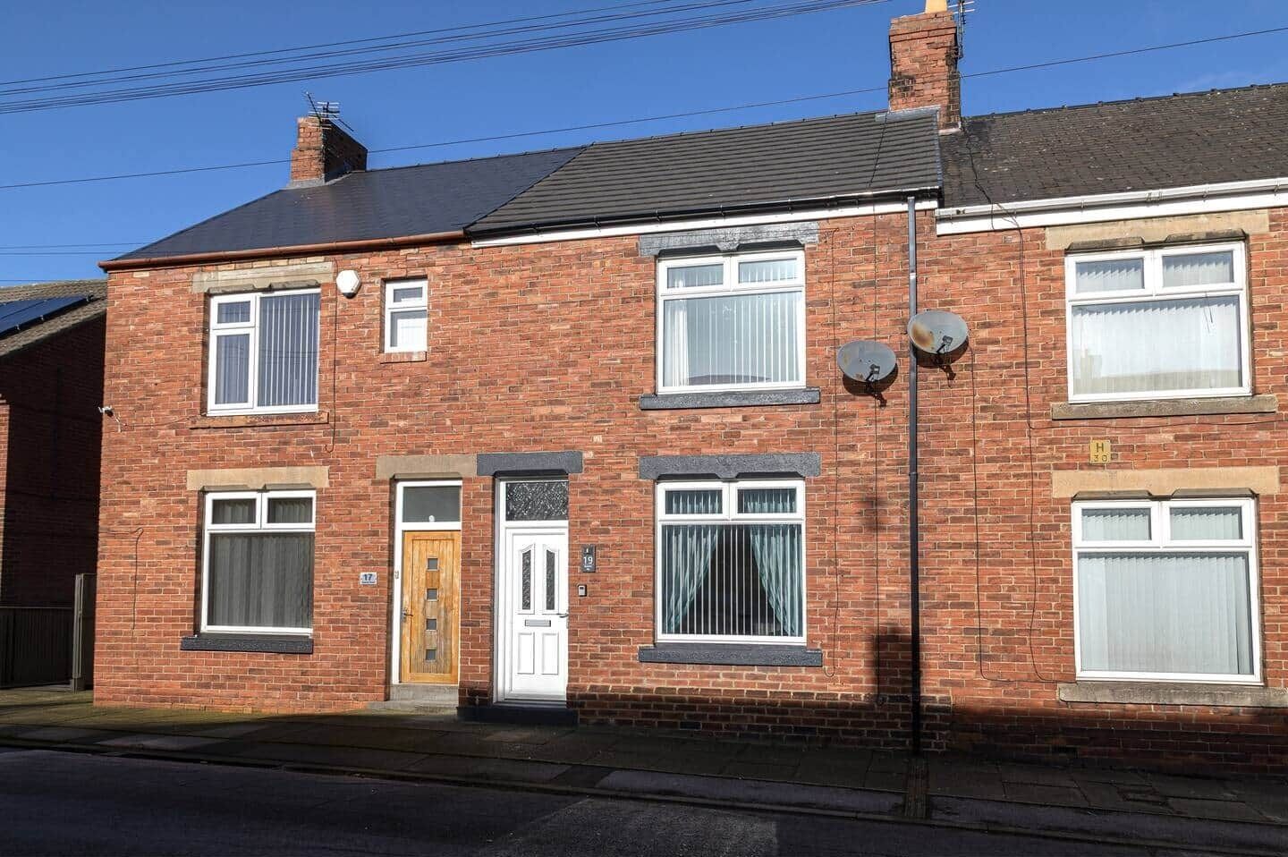 Une maison à deux étages, construite en briques, avec une porte d’entrée, deux fenêtres au rez-de-chaussée et une cheminée.