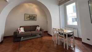 A living room with a sofa, a dining table with chairs, a decorative light fixture, and a window with a view of buildings. - PAOLINA HOME IN ROME (Roma)