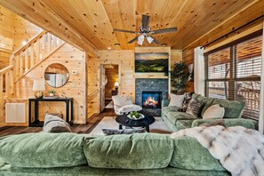 A cozy log cabin interior with a fireplace, green upholstered furniture, and a wooden staircase. - Smoky Mtn Retreat | Sauna + Hot Tub + Game Room (Pigeon Forge)