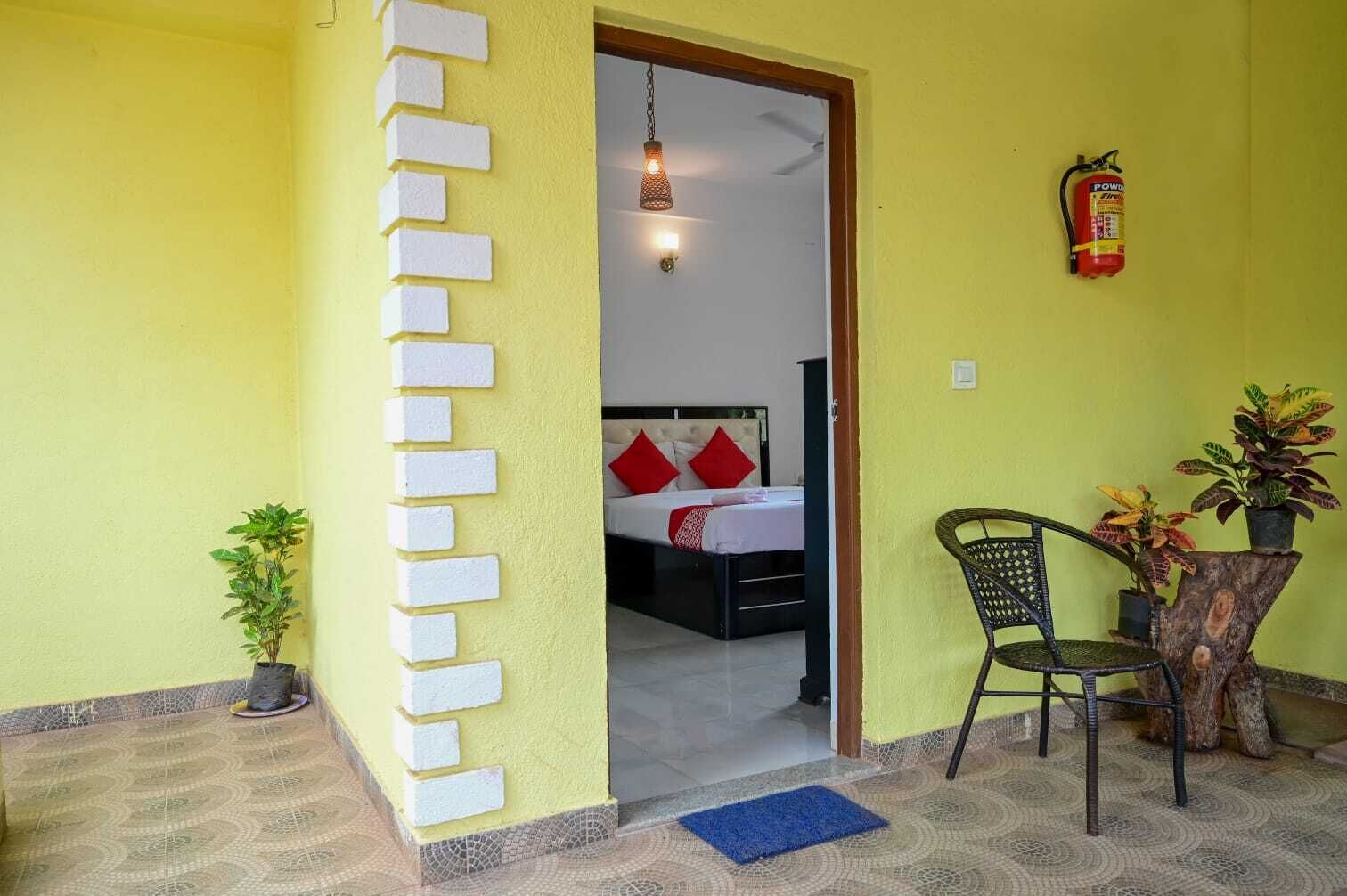A room with a bed, a chair, a small table, a fire extinguisher, and a potted plant.