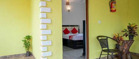 A room with a bed, a chair, a small table, a fire extinguisher, and a potted plant.