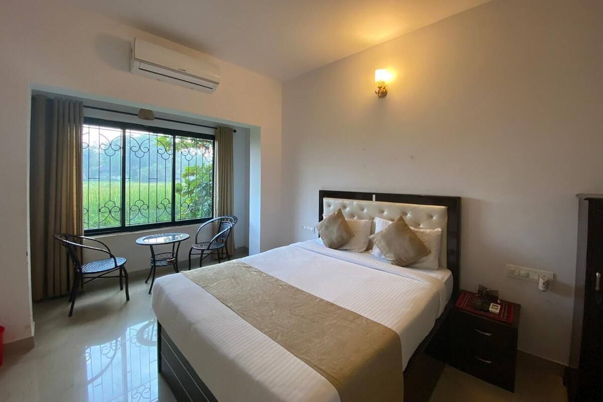 Quarto de hotel com uma cama, uma mesa pequena, duas cadeiras, uma janela com cortinas e um ar condicionado.