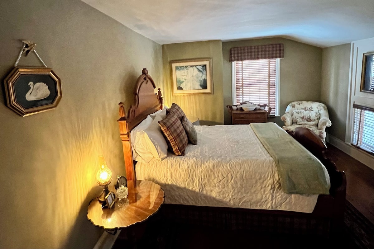 Um quarto bem arrumado com uma cama grande, mesa de cabeceira com abajur, uma cadeira e janela com persianas.