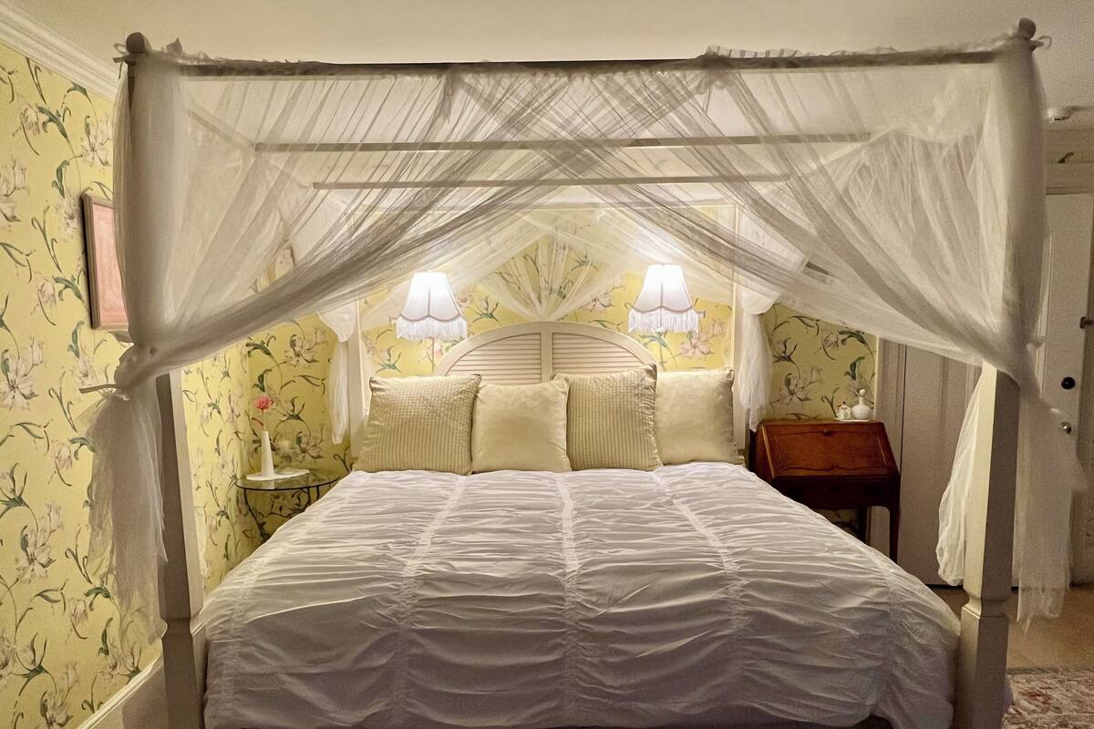 Uma cama de dossel com dossel, com colcha branca, duas luminárias de apoio e papel de parede floral.