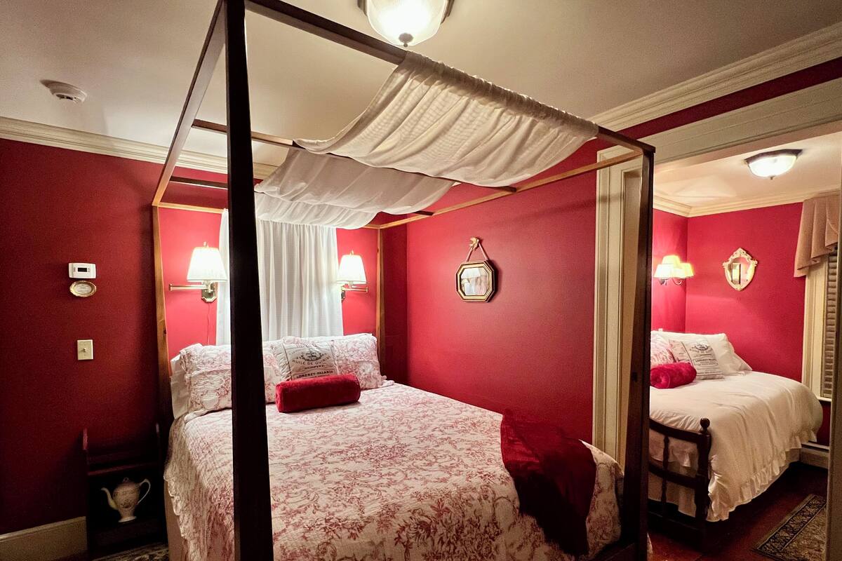 Uma cama com dossel e cortina, uma mesa de cabeceira com abajur, e um espelho na parede.