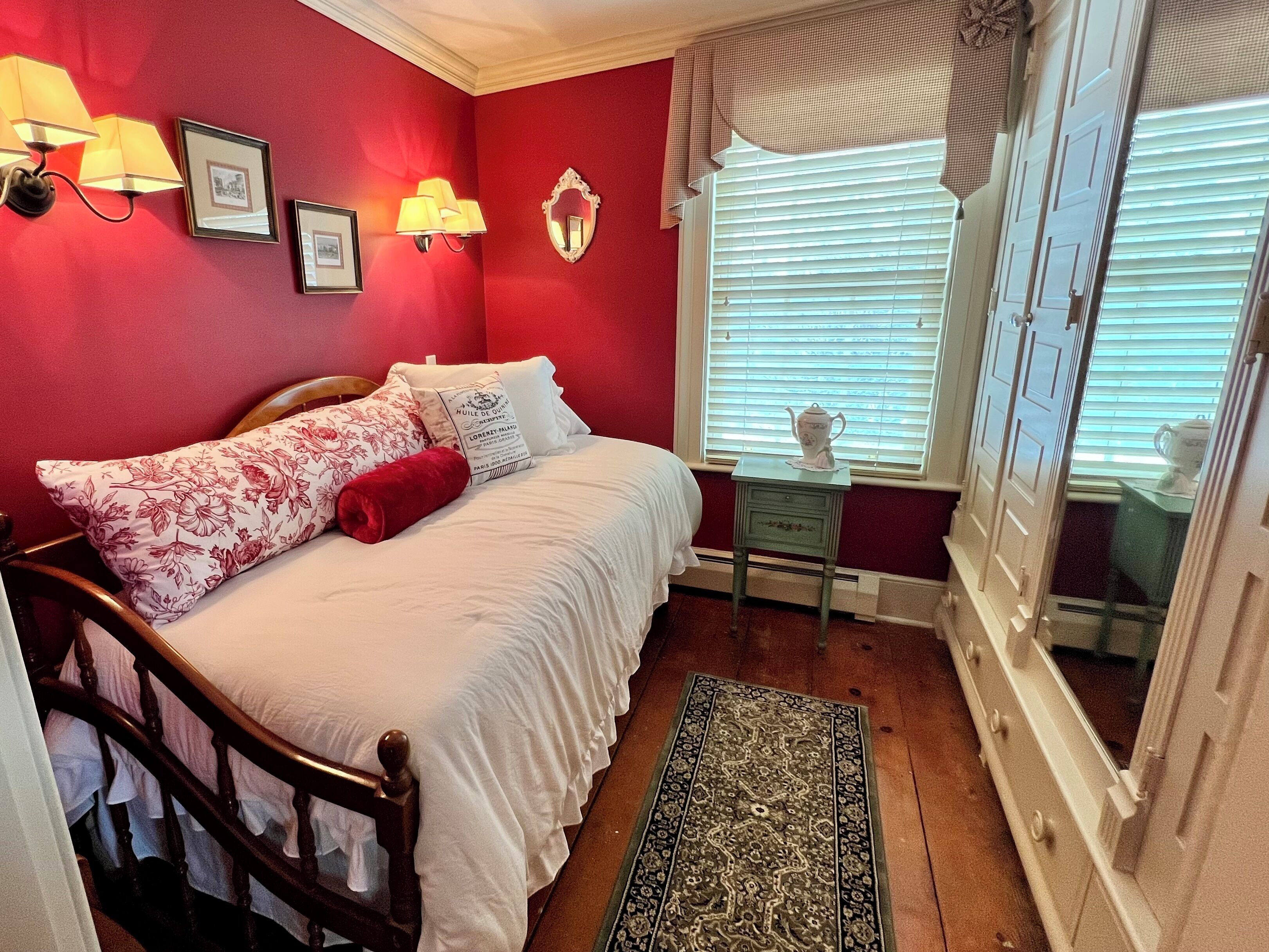 Une chambre à coucher avec un lit en bois, une table de chevet, une fenêtre avec des stores et une petite table sur laquelle se trouve une théière.