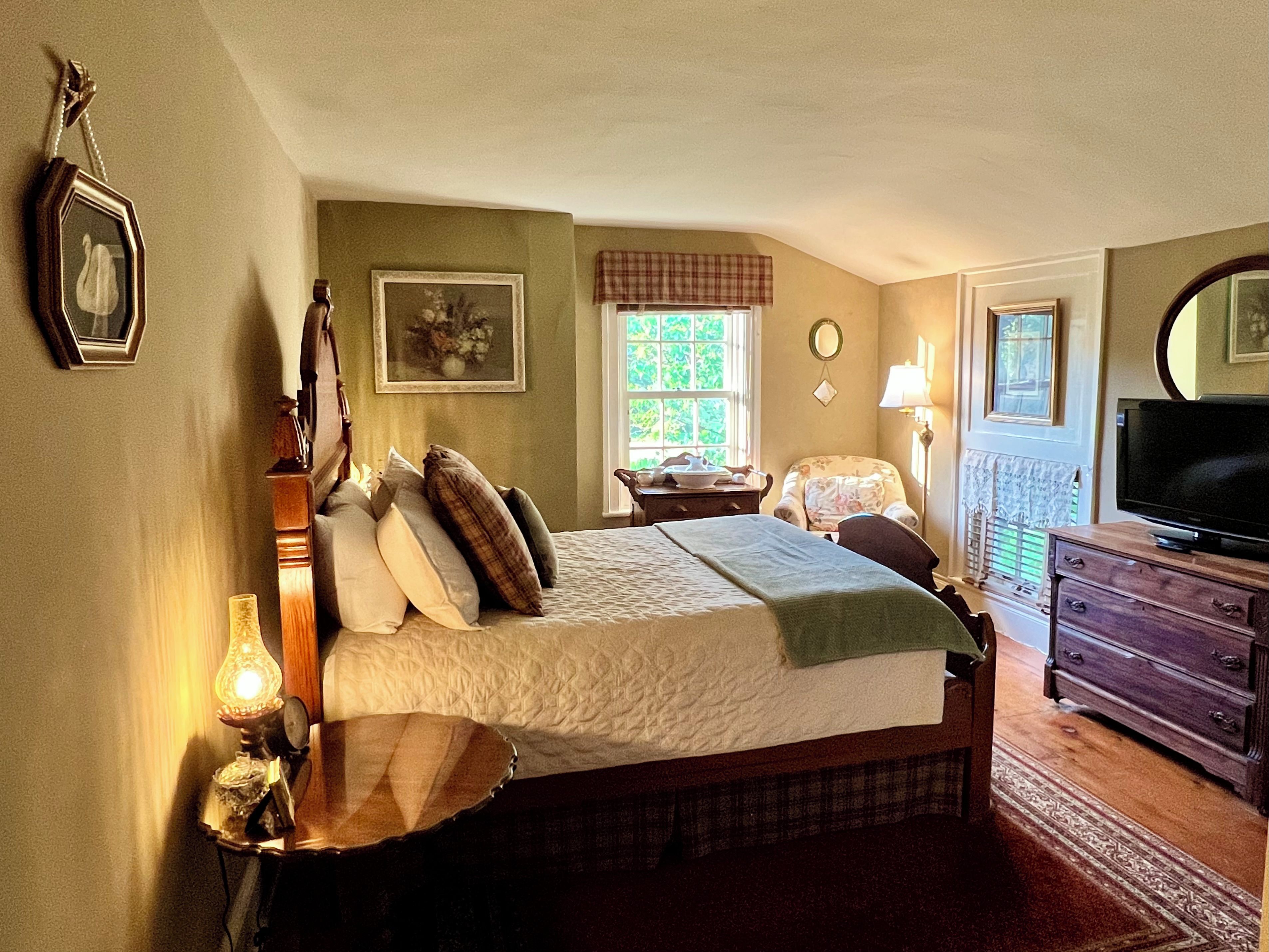 A bedroom with a bed, bedside table, lamp, dresser, television, and a window with curtains.