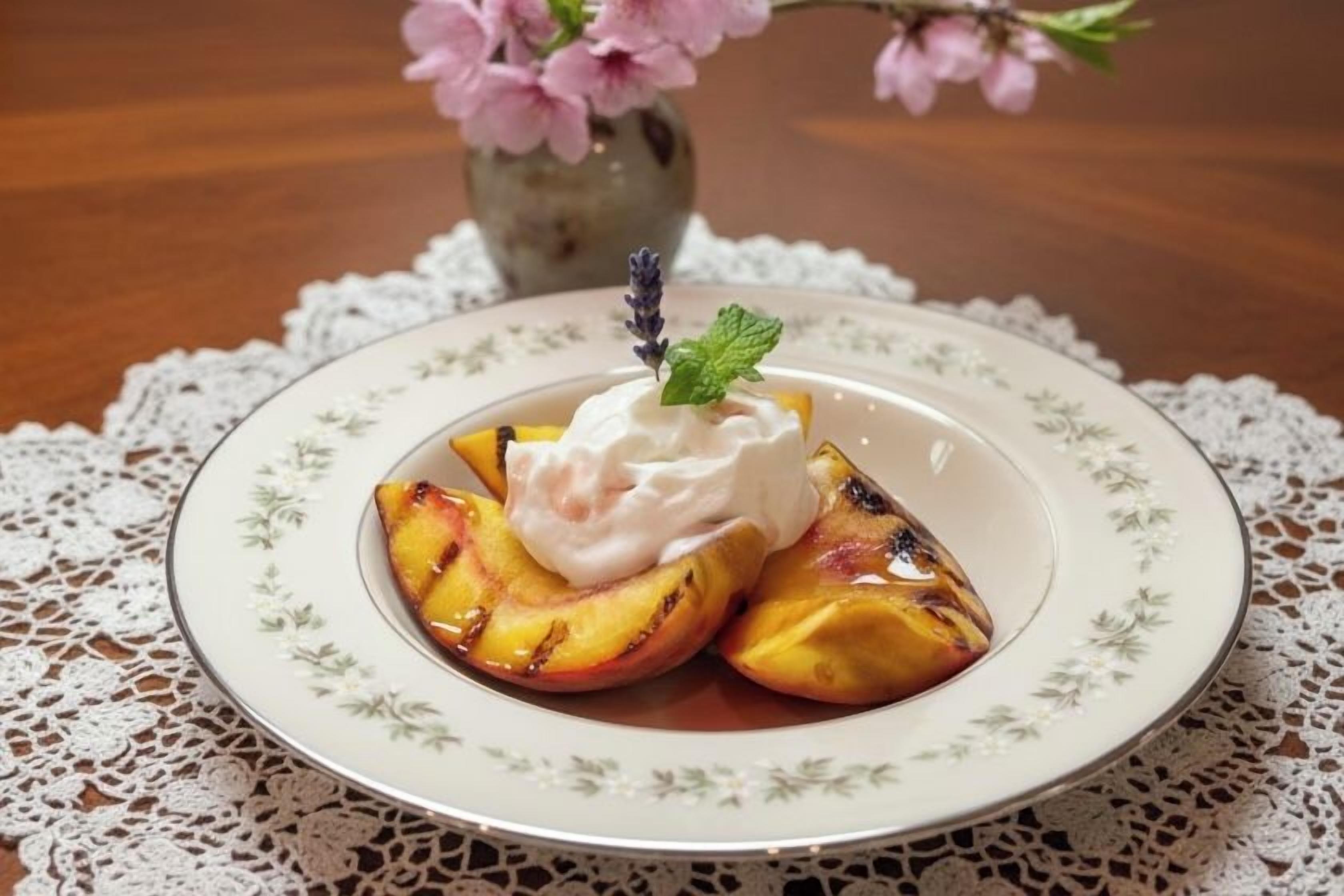 Uma travessa de pêssegos grelhados com chantilly e uma folha de hortelã sobre uma toalha de renda.