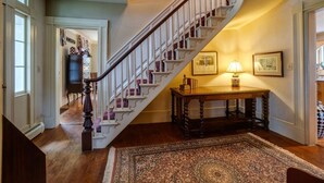 A grand staircase with a white railing and purple accents, leading to a room with a wooden table and a lamp.